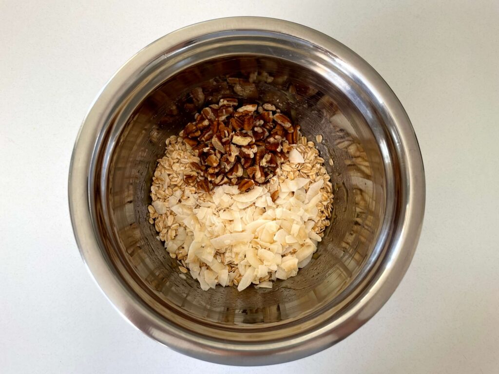 Rolled oats, coconut flakes, and pecans in a stainless-steel bowl