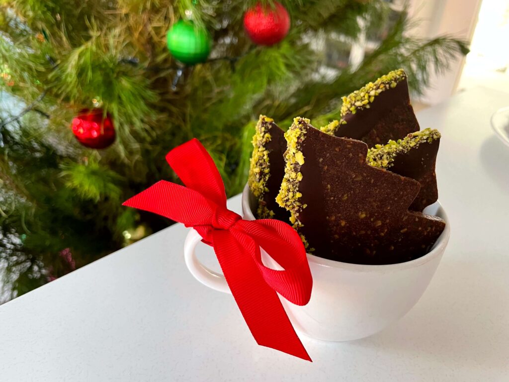 Chocolate pistachio Christmas tree cookies in a cup with a festive red bow and a decorated Christmas tree in the background