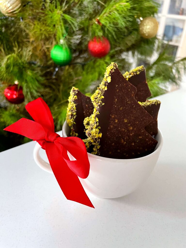 Chocolate pistachio Christmas tree cookies in a cup with a festive red bow and a decorated Christmas tree in the background