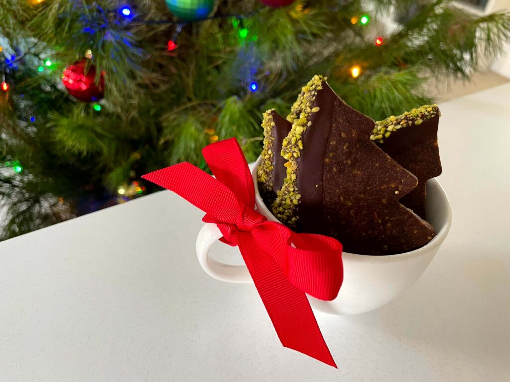 Chocolate pistachio Christmas tree cookies in a cup with a festive red bow and a decorated Christmas tree in the background