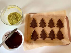 Chocolate pistachio Christmas tree cookies with a bowl of melted chocolate and a bowl of ground pistachios for dipping