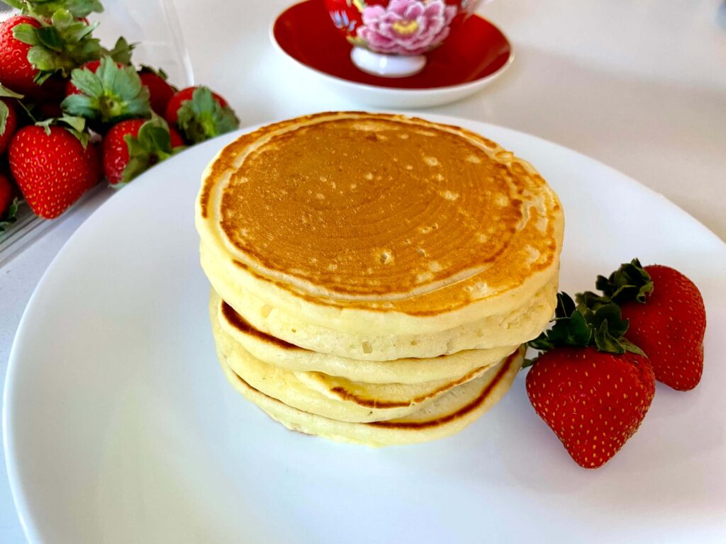 A stack of buttermilk pancakes with strawberries and a teacup in the background