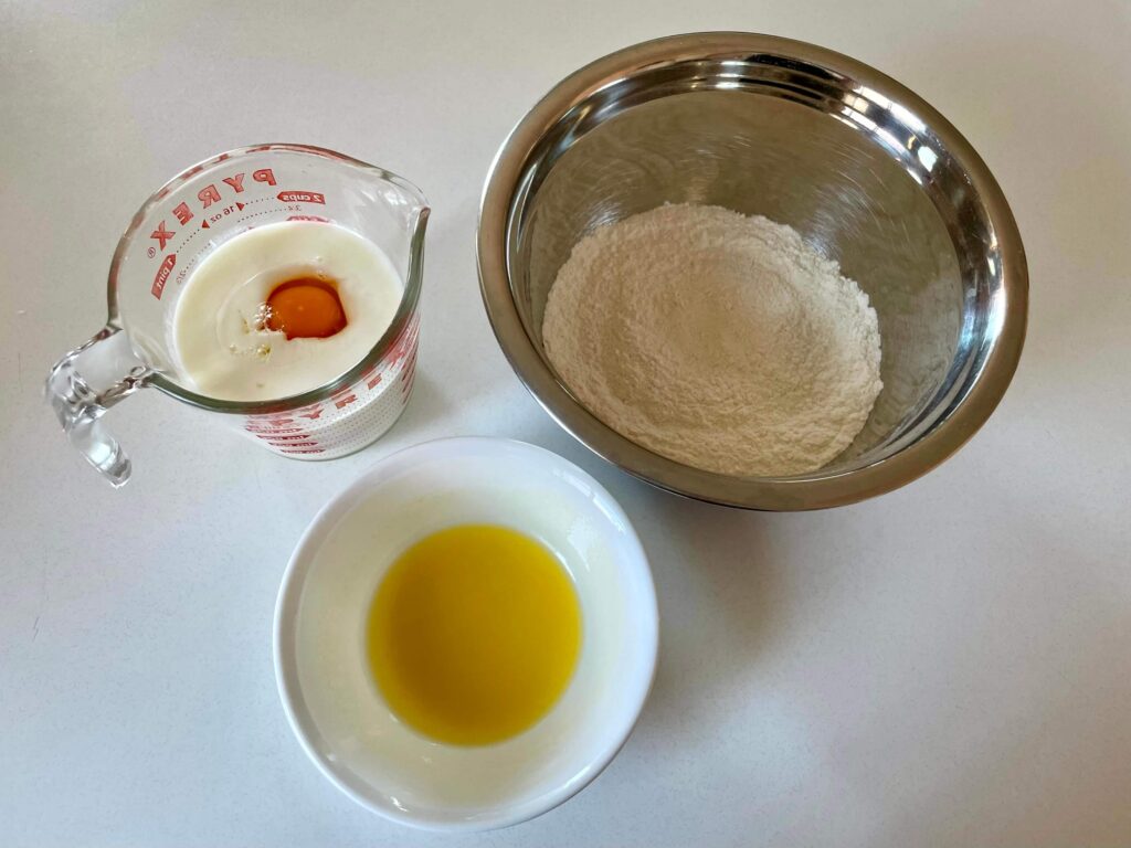 Pancake ingredients: a mixture of flour, sugar, baking powder, and salt; eggs in buttermilk; and a bowl of melted butter.